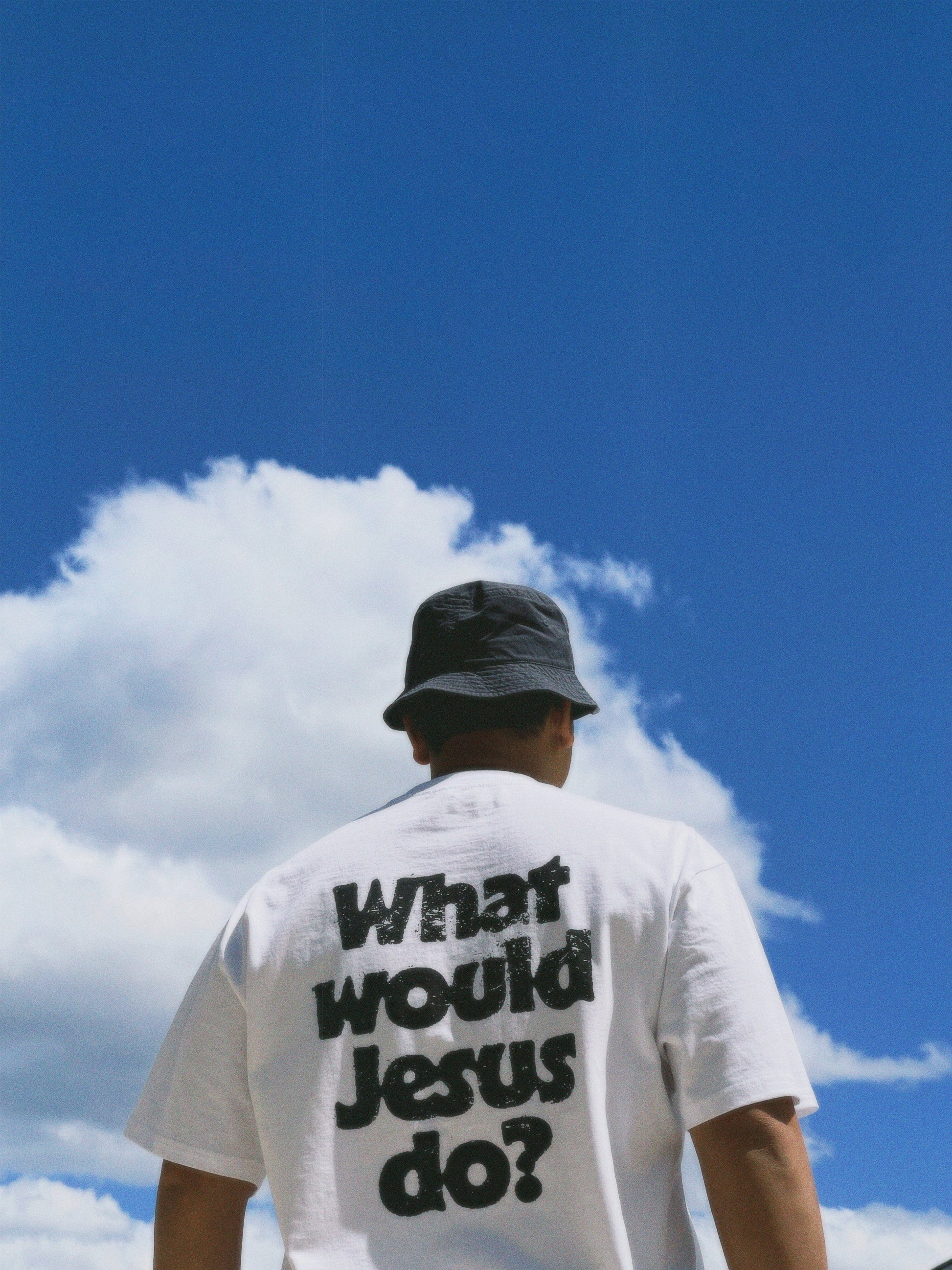 Person wearing a white t-shirt with 'What would Jesus do?' text, standing against a blue sky with clouds.